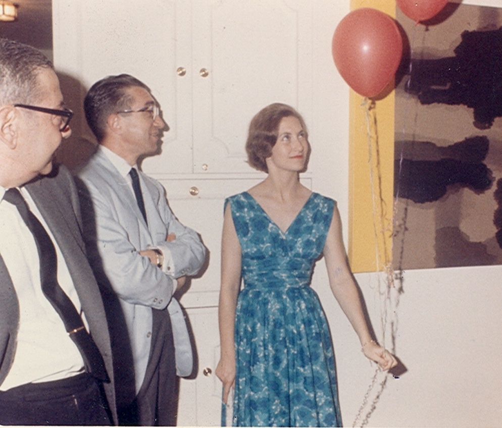 Three adults stand indoors; one woman in a blue dress holds red balloons, while two men in suits stand nearby, smiling.
