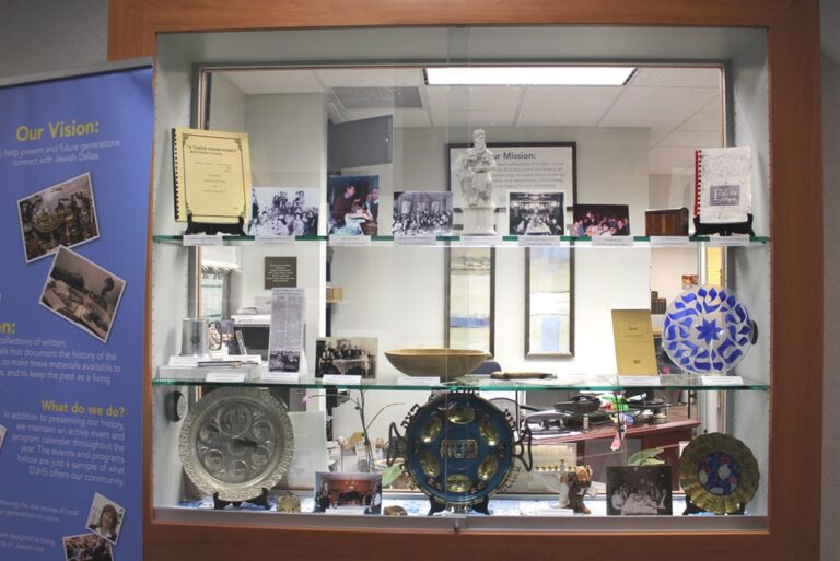 A glass display case with various artifacts, books, photos, and decorative plates. Office space is visible behind the case.