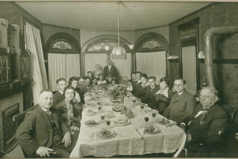 Large group of people in formal attire seated around a dining table set for a meal in a vintage room.