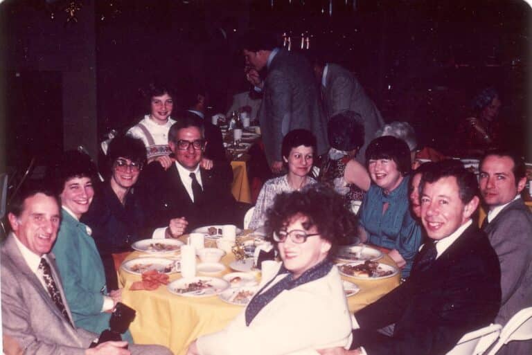 A group of people sit around a dinner table, smiling at the camera during a social gathering.