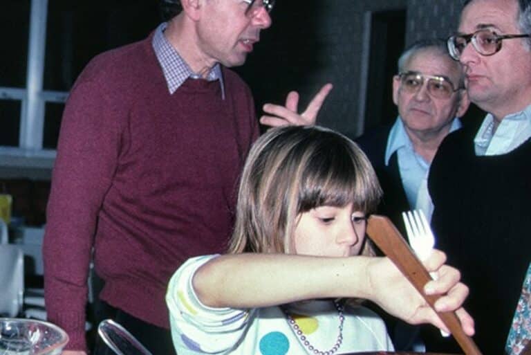 A young girl stirs food with adults standing around her, one gesturing with his hand. Indoor setting.