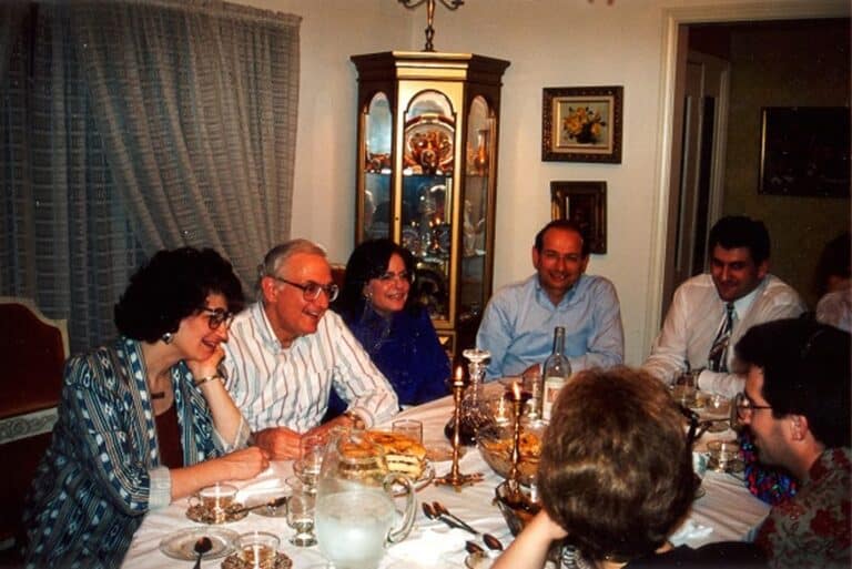 Seven people sit around a dinner table, smiling and talking, with food and drinks in a warmly lit dining room.