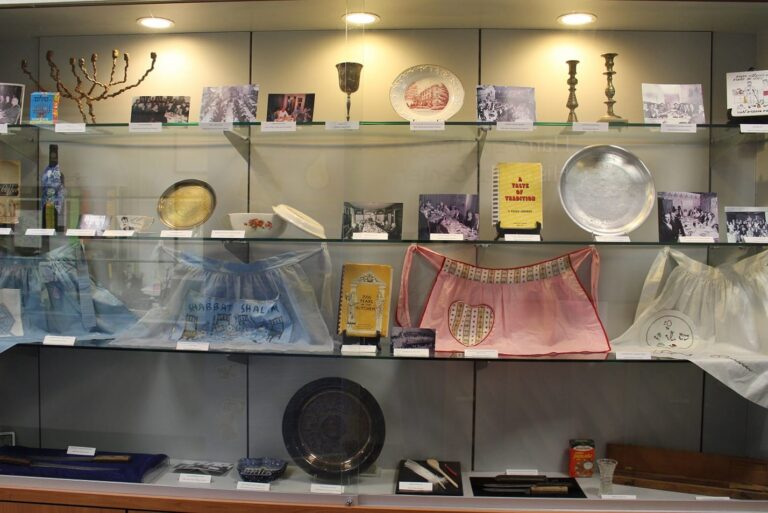 A display case with Jewish ceremonial objects, photos, books, candlesticks, plates, and decorated fabric aprons.