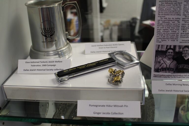 Glass tankard, black bottle opener, and gold pin on display with descriptive labels in a glass case.