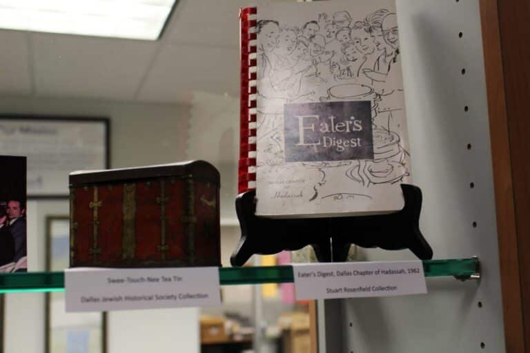 A vintage tin and a spiral-bound cookbook titled “Eater’s Digest” are displayed on a glass shelf in a museum.