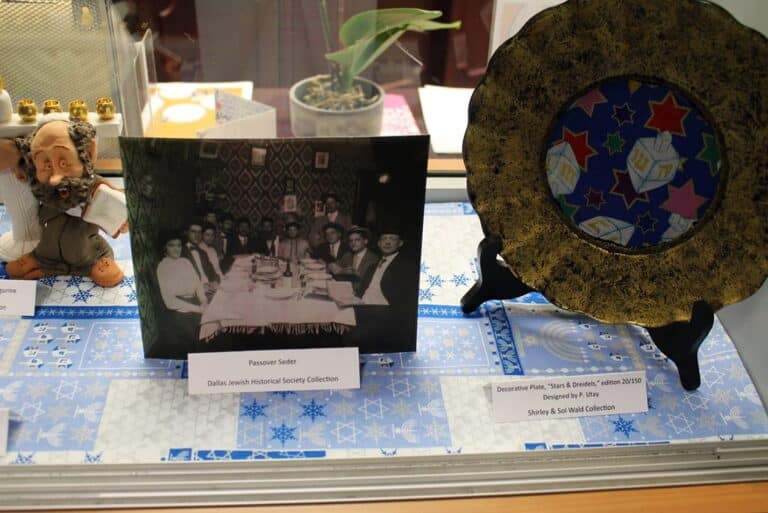 Display case with a photo of a Passover Seder, a decorative plate, and a figurine on a Star of David table mat.