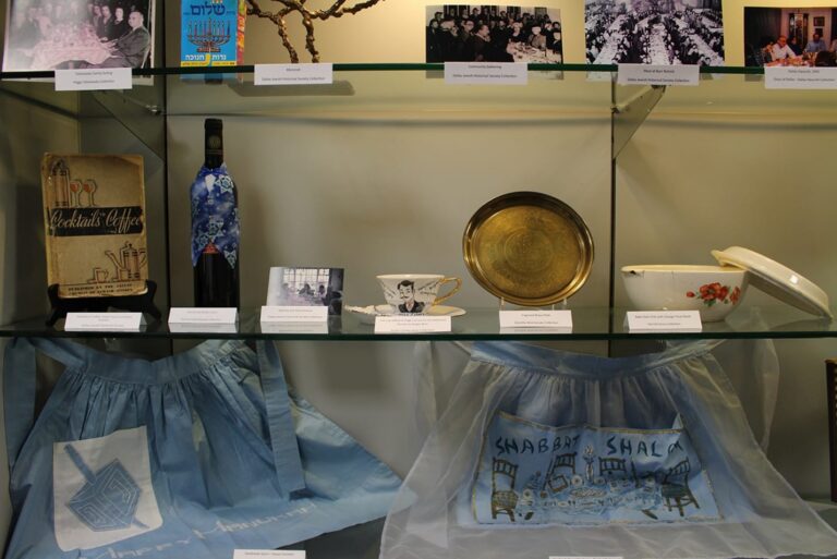 Museum display case with vintage kitchen items, books, aprons, photos, and a brass plate on glass shelves.