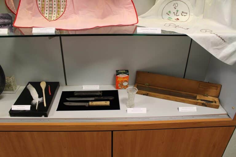 Museum display with a feather pen, two knives, a glass, a juice box, and a long wooden case on a shelf.