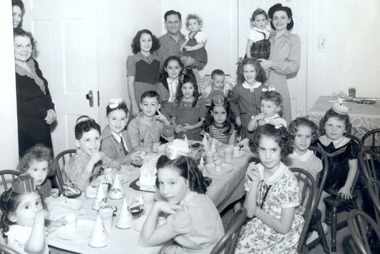 A black-and-white photo of children at a birthday party, with adults standing behind them, smiling at the camera.