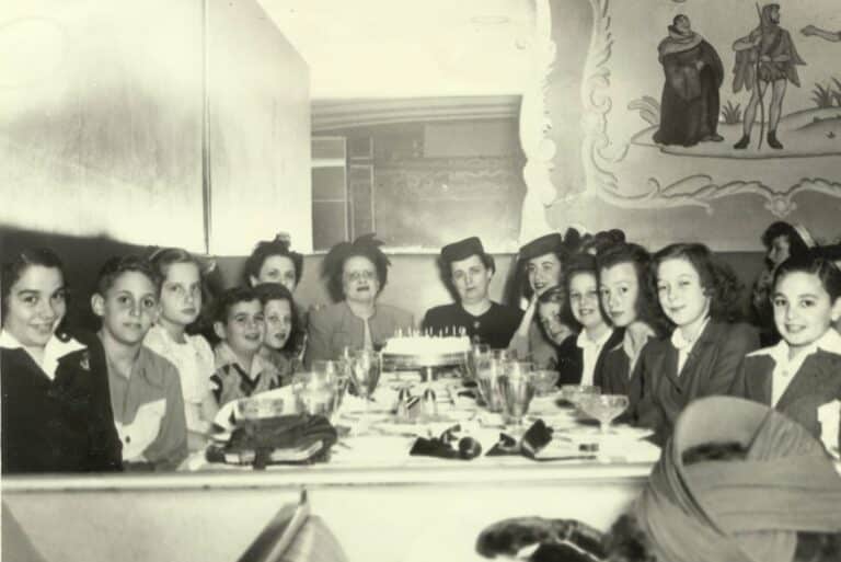 Black and white photo of children and adults around a table with a cake, smiling at a celebration.
