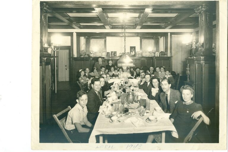 A large family gathers around a long table for a meal in a wood-paneled dining room, April 1943.