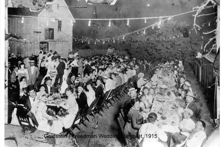 Large outdoor wedding banquet at night, 1915, with many guests seated at long tables under string lights.