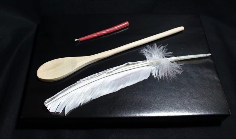 A red candle, a wooden spoon, and a white feather lie on a glossy black surface against a dark background.