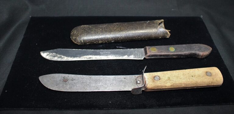 Two old knives with worn blades and wooden handles, lying next to a weathered leather sheath on a black surface.