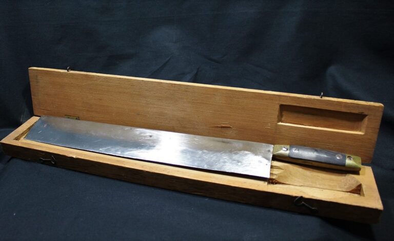 A large knife with a wooden handle rests in an open wooden box against a black background.