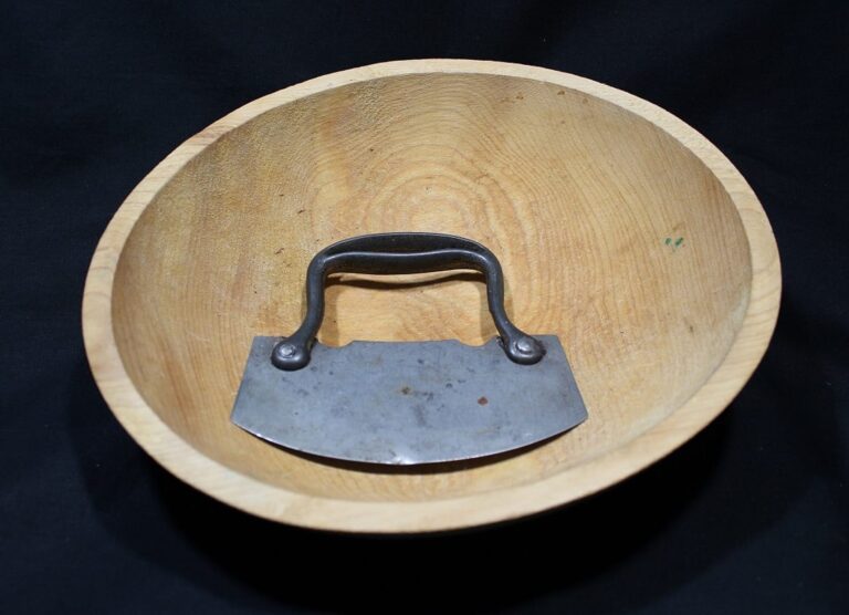 A wooden bowl with a metal mezzaluna chopper inside, both placed against a black background.