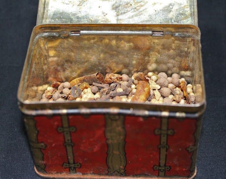 An open tin box with a red design holds a mix of dried spices and herbs on a dark surface.