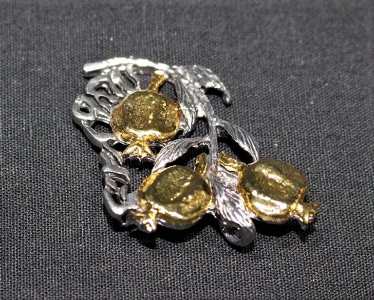 A silver and gold brooch shaped like a branch with three pomegranates, resting on a black fabric background.
