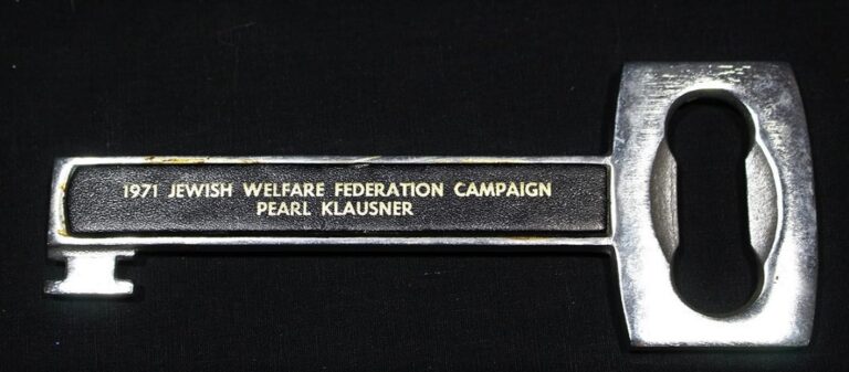 A metal key with “1971 Jewish Welfare Federation Campaign Pearl Klausner” engraved on it, against a black background.