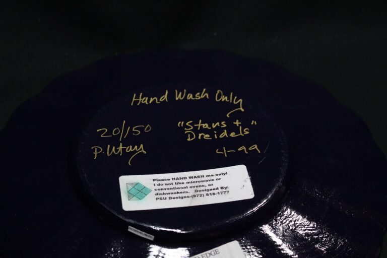 A dark plate’s underside with handwritten notes and a sticker reading “Hand wash only; no microwave or conventional ovens.”.