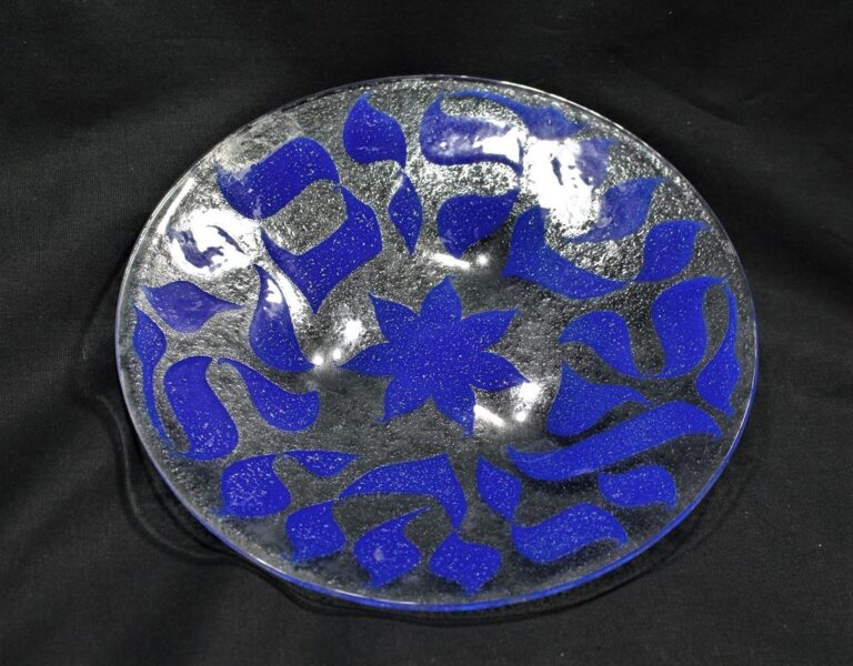 A round glass plate with blue abstract shapes and a blue floral pattern in the center on a black background.