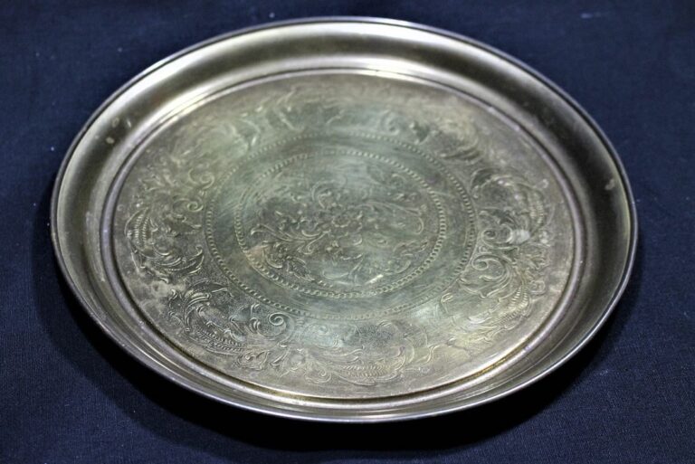 A round, ornate metal plate with intricate engraved patterns, placed on a dark cloth background.