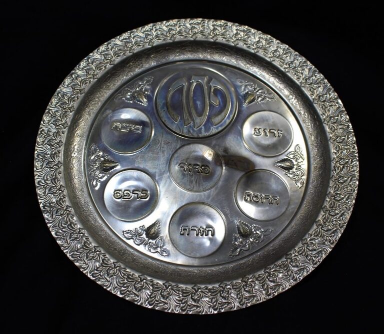 Silver Passover Seder plate with Hebrew labels for ritual foods, ornate floral border, on a dark background.