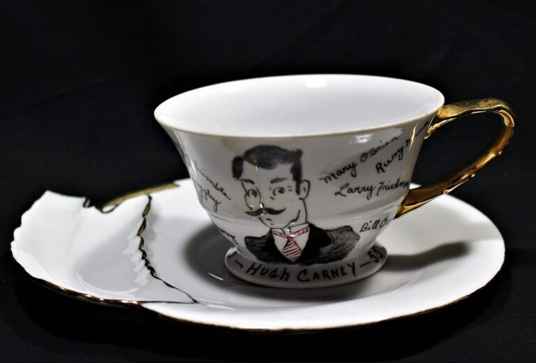 Teacup with gold handle and matching saucer, featuring sketch of a man and signatures, against a black background.