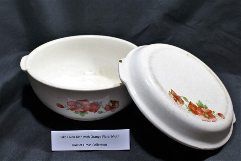 White bake oven dish with orange floral design, lid beside it, on black fabric. Label describes floral motif and the collection.