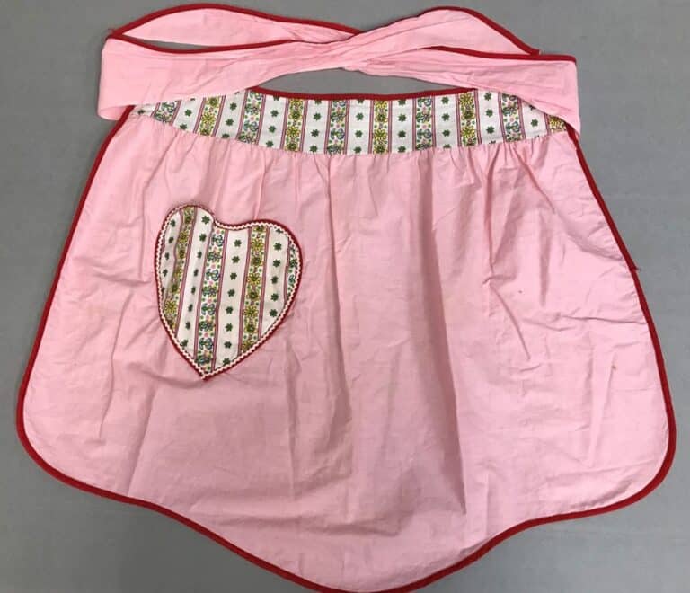 Pink apron with a heart-shaped pocket and floral trim, red edging, and a waistband with matching floral pattern.