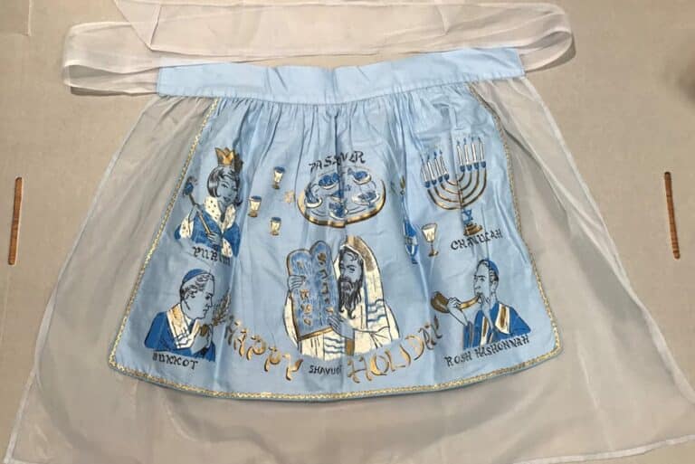Light blue apron with Jewish holiday symbols, including menorahs, Torah, and portraits, with “Happy Holiday” text embroidered.