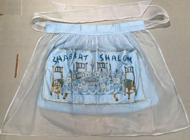 A sheer apron with “Shabbat Shalom” and a table set for Shabbat embroidered on the front panel.