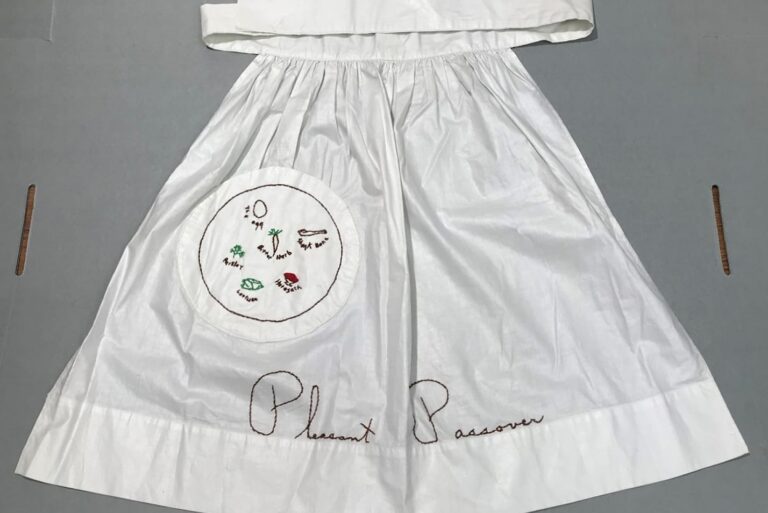White apron with a Passover Seder plate design and “Pleasant Passover” embroidered in brown thread at the bottom.