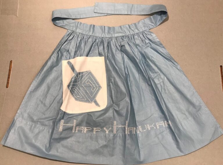 Light blue apron with “Happy Hanukkah” and a dreidel design on the front, displayed on a flat surface.