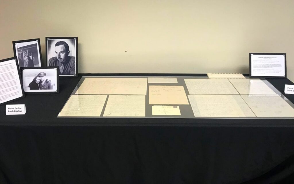 Display table with framed photos, documents, and papers under glass; two signs and a descriptive placard are visible.