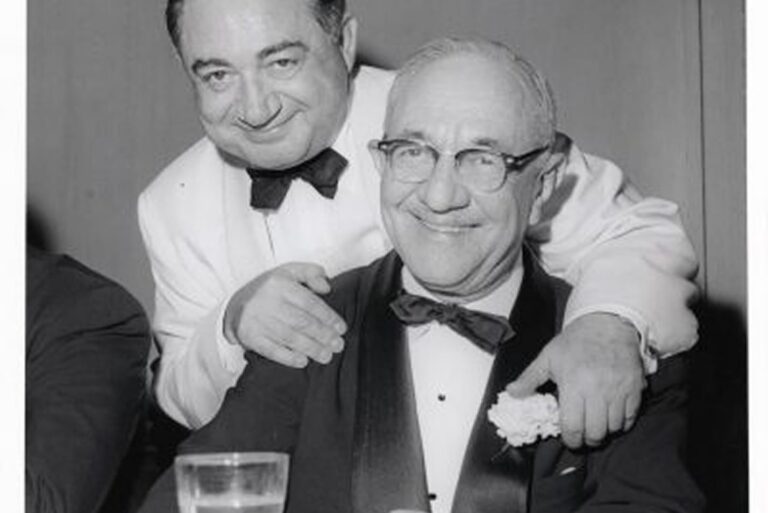 Two older men in tuxedos smiling at a formal event, one standing with his hand on the other’s shoulder.