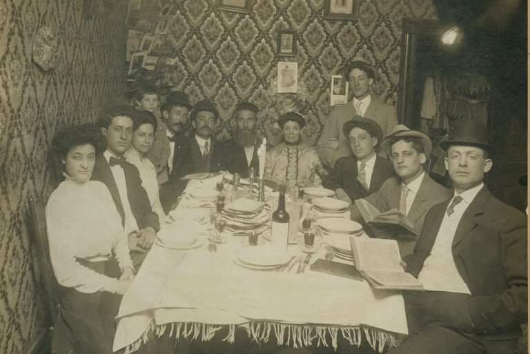 Twelve people in early 1900s attire sit around a dining table set for a meal in a patterned, vintage room.