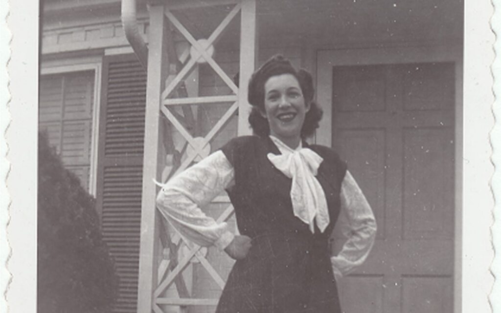 Smiling woman in vintage clothing stands in front of a house with hands on hips, black-and-white photo.