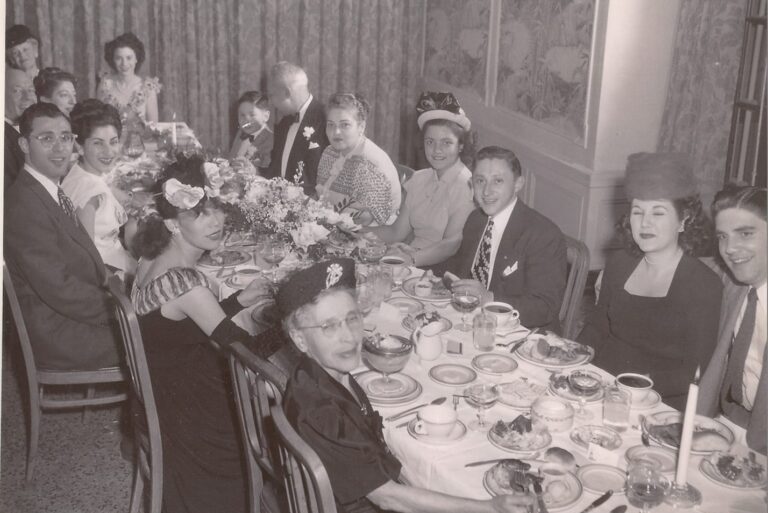 Group of people dressed formally, sitting around a table set for a meal, smiling and talking at a gathering.