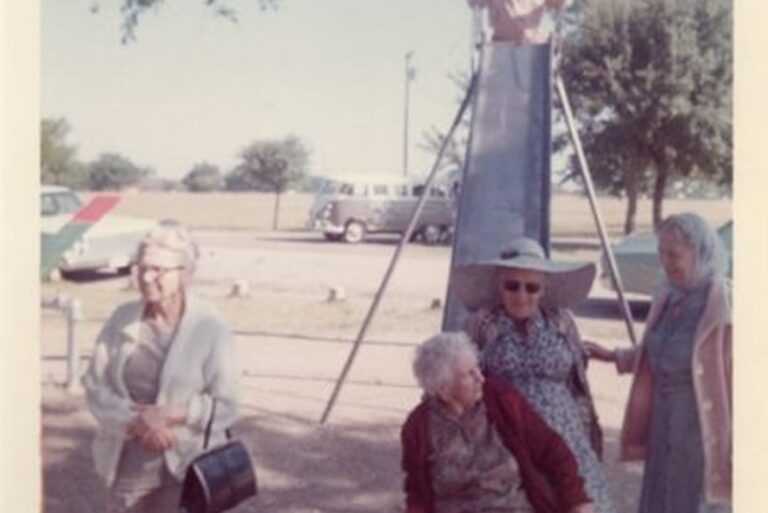 Four elderly women stand and sit near a playground slide with a child at the top, outdoors on a sunny day.