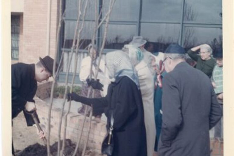A group of people gathers outside a building as a man shovels soil around a newly planted tree.