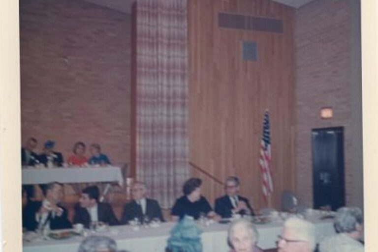 People sit at banquet tables in a wood-paneled room with an American flag and brick walls in the background.