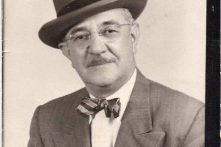 Older man in a suit, bow tie, and hat, with glasses and a mustache, posing for a portrait, black-and-white photo.