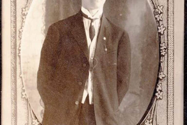Sepia photo of a man in a suit and tie, standing with one hand in his pocket, in an ornate oval frame.