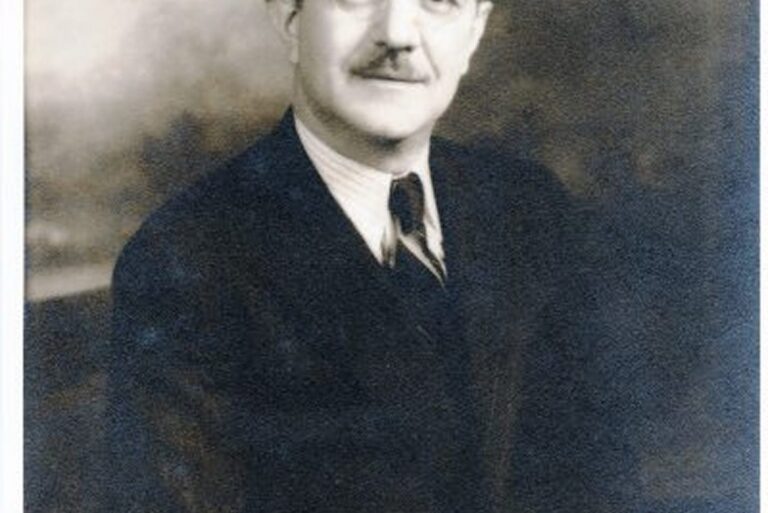 A man with a mustache in a suit and tie poses for a formal black-and-white portrait, looking slightly to the side.