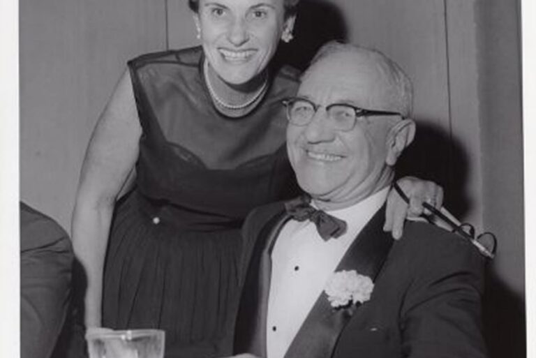 A smiling woman stands behind a seated man in a tuxedo holding a drink at a formal event.
