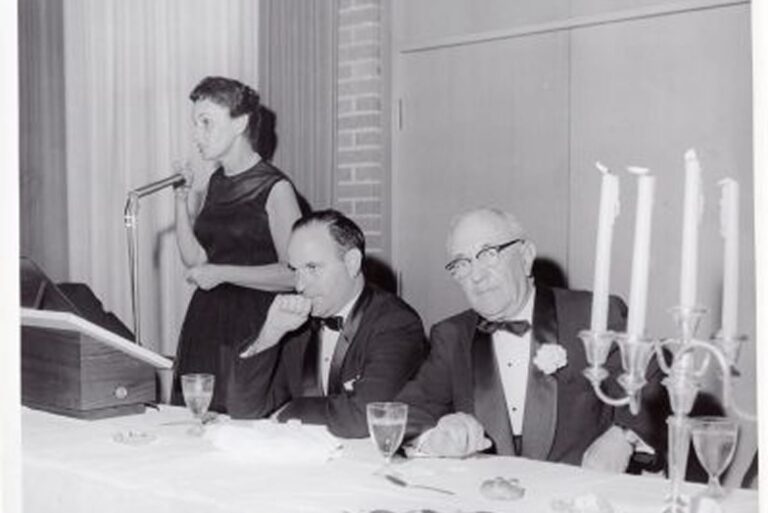 A woman speaks at a podium while two men in tuxedos sit at a table with candles and glasses.