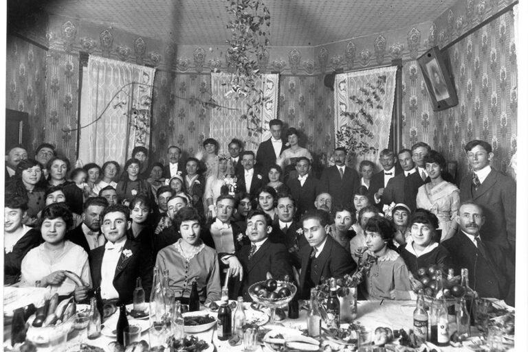 Large group of people in formal attire gathered in a decorated room around tables with food and drinks.