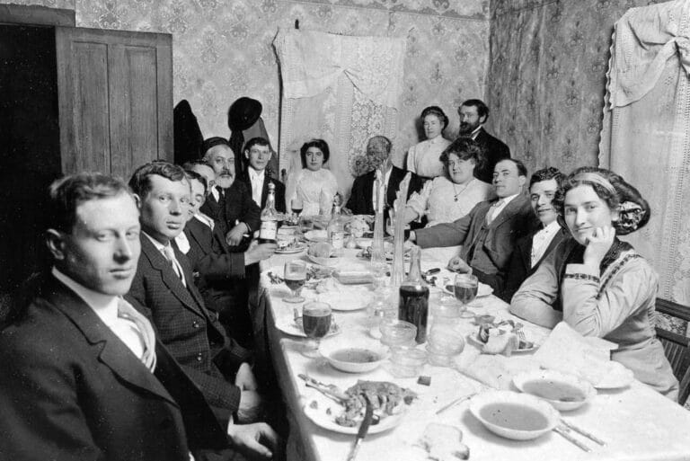 A large group of people dressed formally sit around a dining table set for a meal in a vintage room.
