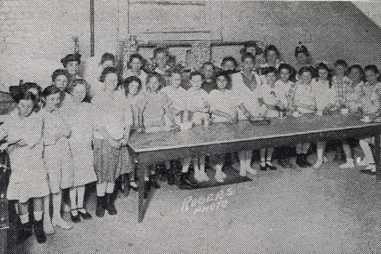 A group of children and a few adults pose together behind a long table in a black and white photo.
